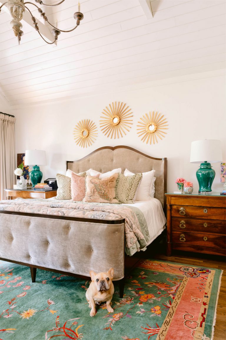 A cozy bedroom features a plush bed with a tufted headboard, adorned with patterned pillows. Above the bed, three sunburst wall decorations provide an artistic touch. A French Bulldog sits on a vibrant rug with floral motifs. Wooden nightstands with green lamps flank the bed.