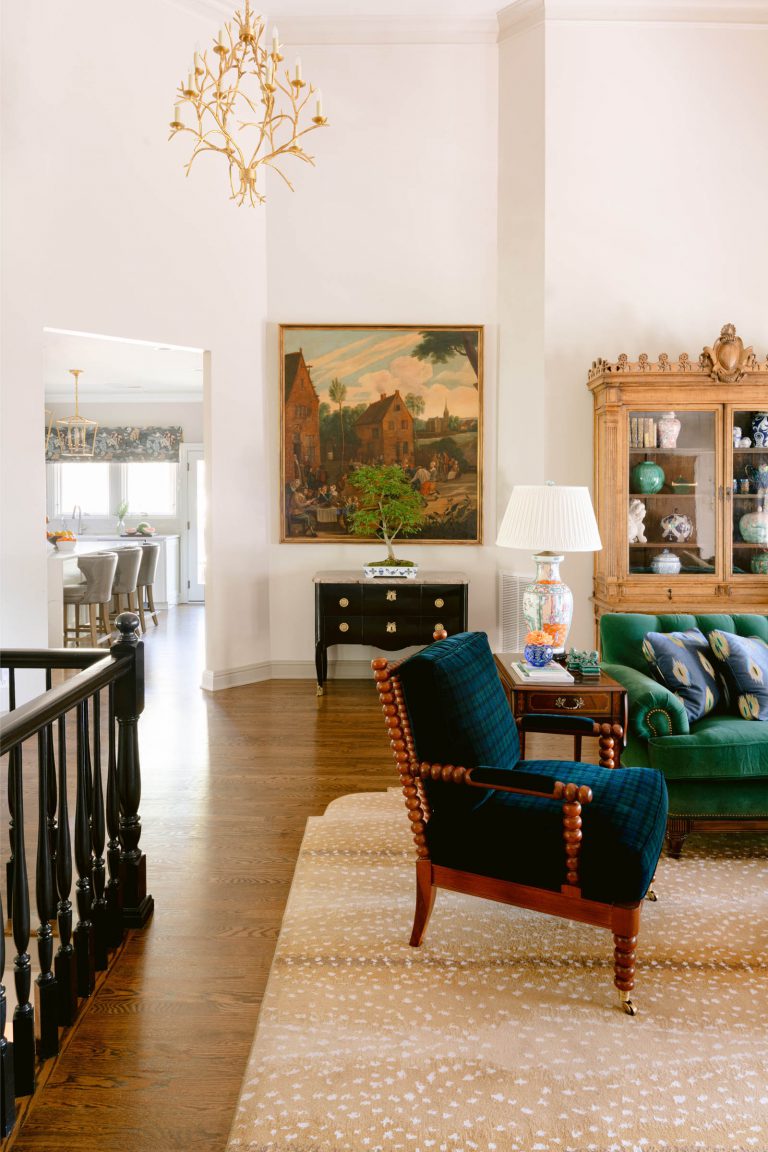 A cozy living room with wooden flooring features a green sofa, a dark checkered armchair, and a wooden cabinet displaying dishes. A large framed painting hangs above a small black chest with a potted plant. A gold chandelier hangs from the ceiling, illuminating the space.