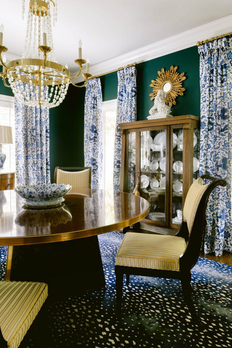 A dining room with a polished wooden table, striped yellow upholstered chairs, and a large, ornate chandelier. The walls are dark green, adorned with blue-and-white patterned curtains. A glass cabinet filled with white china stands against the wall, illuminated by natural light.