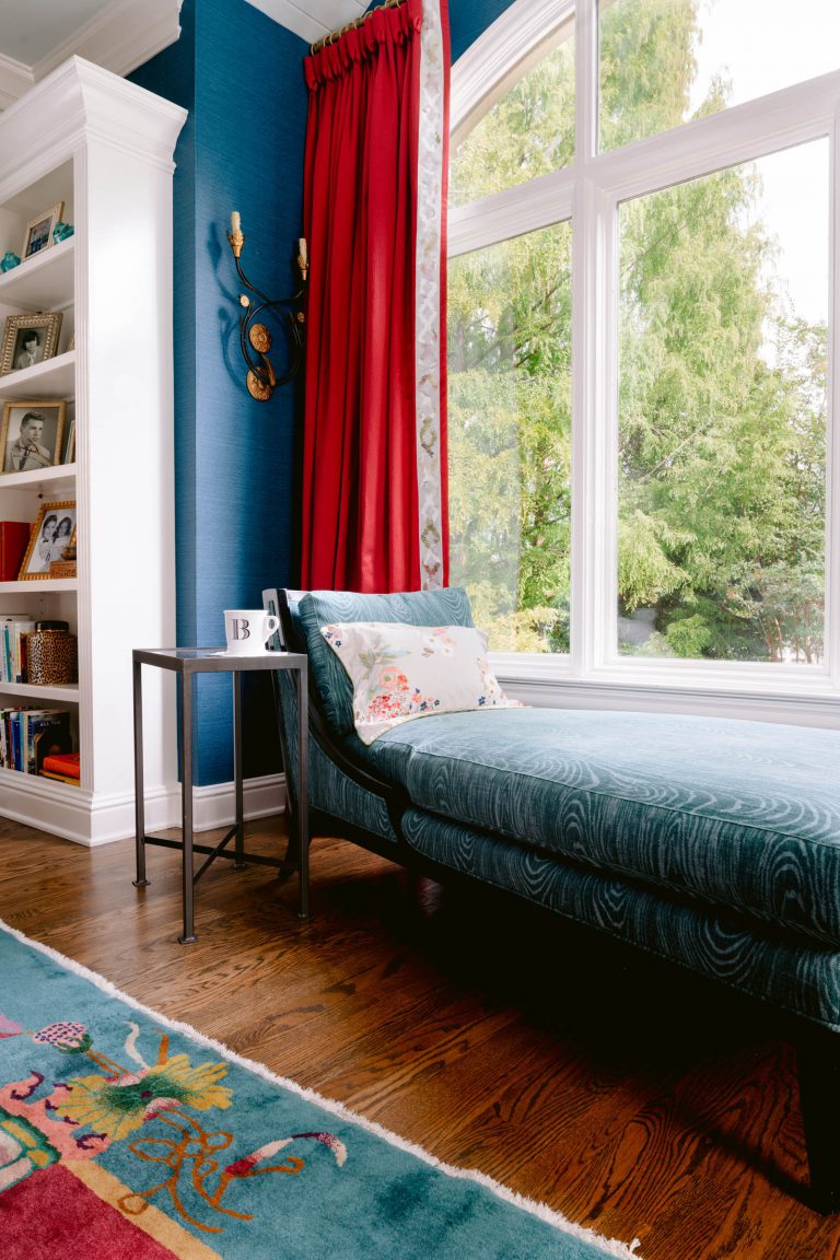 A cozy reading nook features a blue chaise lounge with a floral pillow near large windows with green views. Red curtains add a bold touch. A small side table with a mug rests beside the chaise. Built-in bookshelves display books and framed photos. The wooden floor has a vibrant, patterned rug.