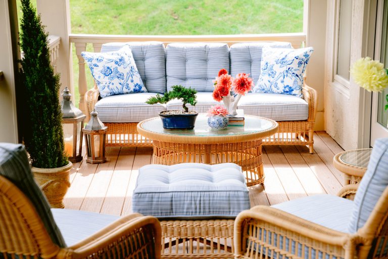 A cozy outdoor patio with wicker furniture, including a sofa, armchairs, and a round glass-topped coffee table. The seats have blue and white cushions. The table is adorned with red flowers and books. A small bonsai adds greenery, while the backdrop shows lush green grass outside.