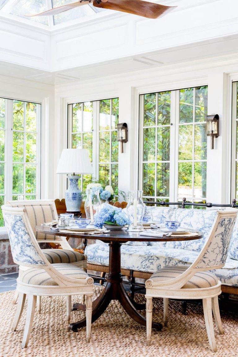 A sunlit dining nook features a round wooden table surrounded by upholstered chairs in blue and white patterns. A center vase displays blue hydrangeas. Large windows reveal lush greenery outside. A woven rug covers the floor, and sconces adorn the walls, beneath a skylight.