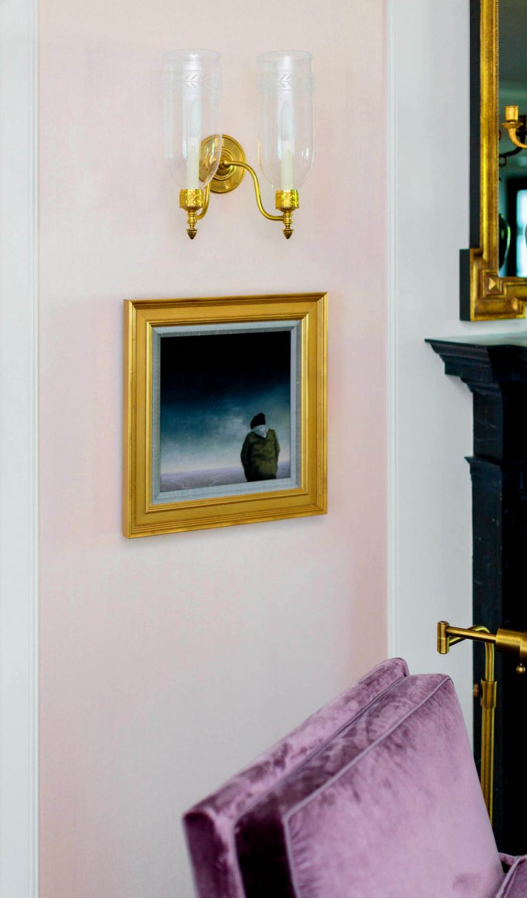 A framed painting with a gold frame hangs on a pale pink wall, depicting a solitary figure in a dark landscape. Above it, a brass wall sconce with two clear glass shades is mounted. In the foreground, a plush purple chair is partially visible. A black fireplace is on the right.