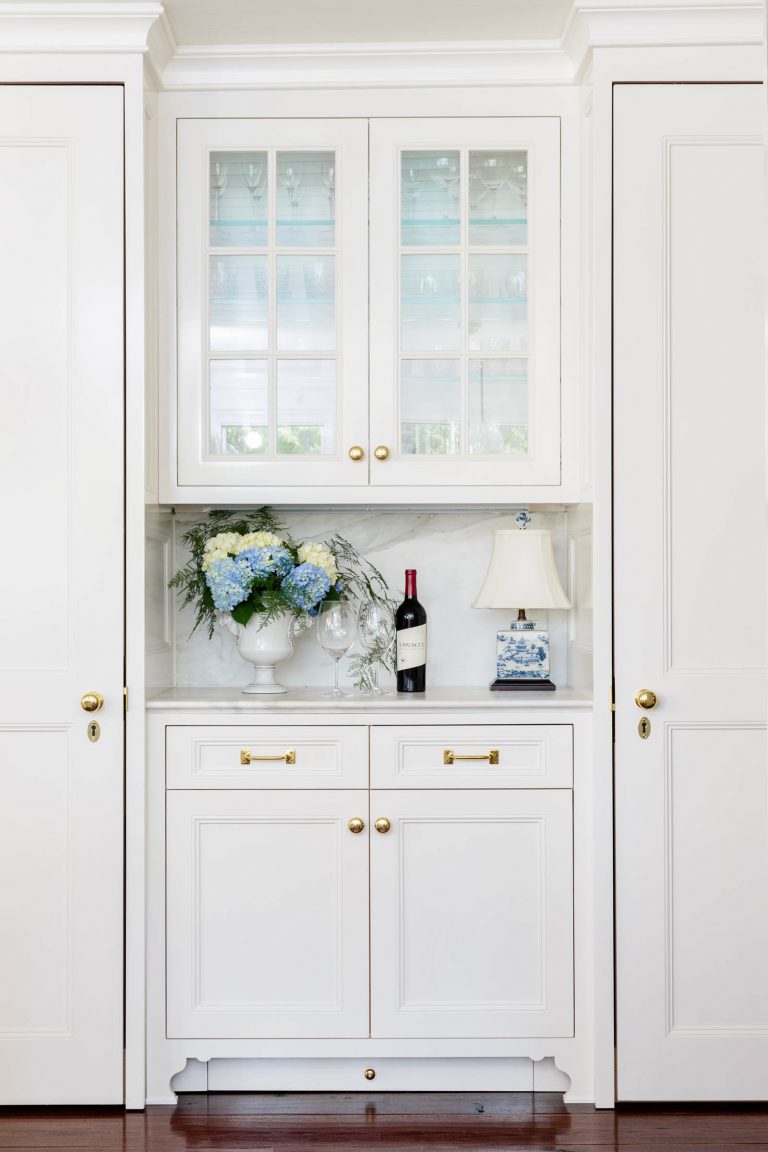 A white cabinet with glass-paneled doors displaying glassware sits against a white wall. Below, a countertop holds a vase with blue and white flowers, a wine bottle, a wine glass, and a small decorative lamp. The cabinet and countertop have gold knobs and handles, with wooden flooring below.