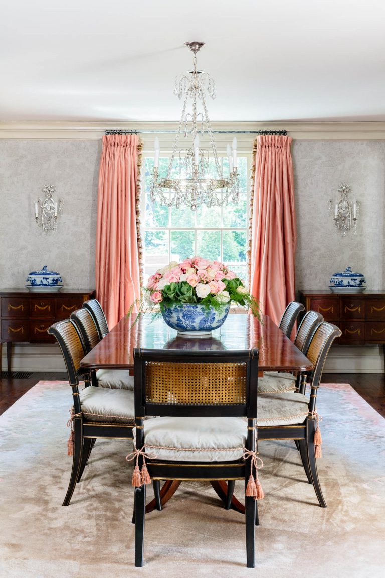 Elegant dining room with a long wooden table surrounded by eight chairs with cushioned seats. A large floral arrangement in a blue vase sits on the table. Crystal chandelier hangs above. Peach-colored drapes frame the window, with blue and white vases on sideboards against light wallpaper.