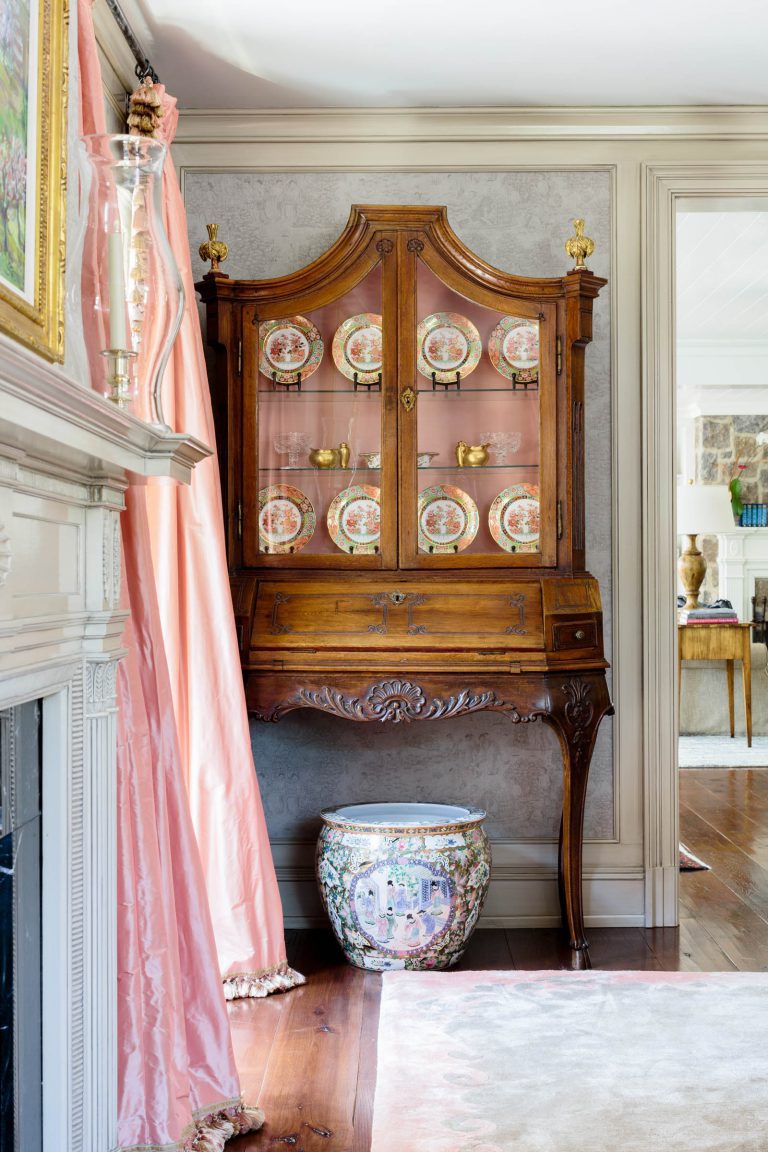 A vintage wooden cabinet with glass doors displays ornate plates and teacups. Below, a decorative ceramic planter with floral designs rests on a wooden floor. Pink curtains hang beside a white mantel. The room features elegant, classic furnishings and soft, natural lighting.