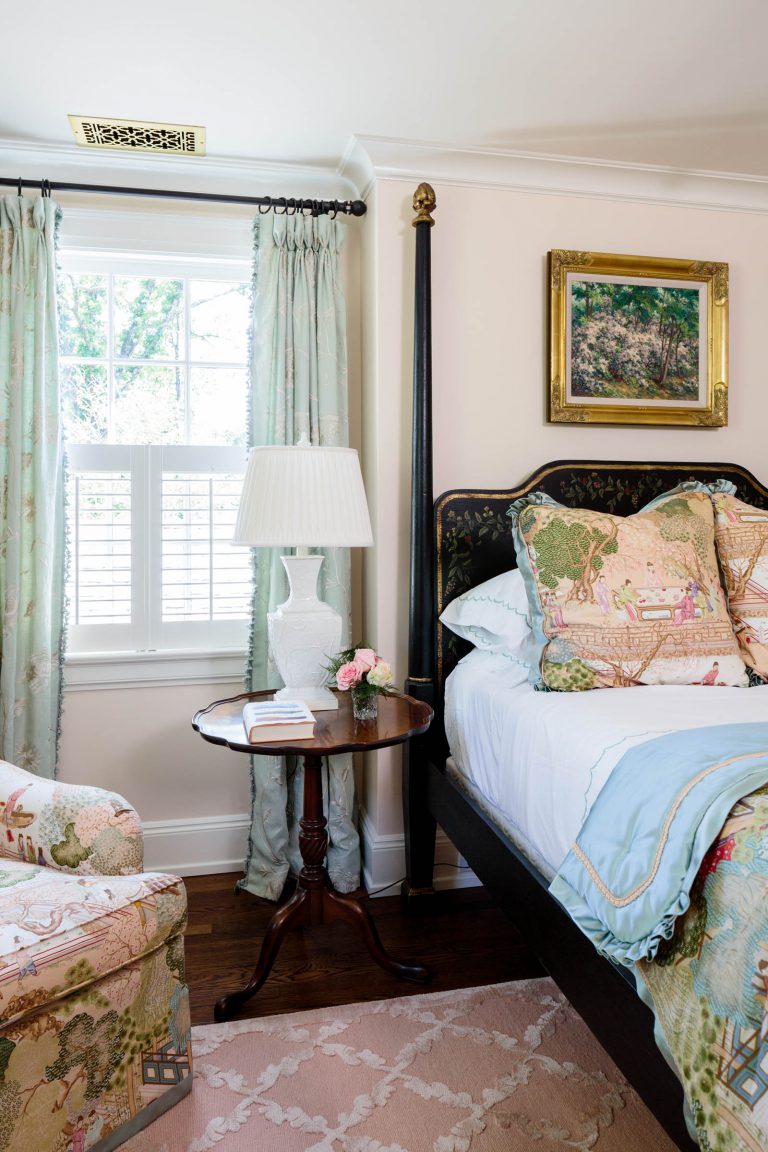 A cozy bedroom features a bed with patterned pillows and a light blue blanket. A wooden nightstand holds a lamp, a book, and a small vase of pink flowers. Near it is a floral armchair. A window with curtains allows natural light in. A framed painting hangs on the cream-colored wall.