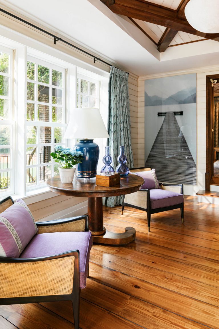 A cozy living room features wooden floors and a vaulted ceiling with exposed beams. Two purple cushioned chairs surround a round wooden table with blue vases, a lamp, and a potted plant. Large windows let in natural light, and a curtain hangs beside a scenic wall mural.