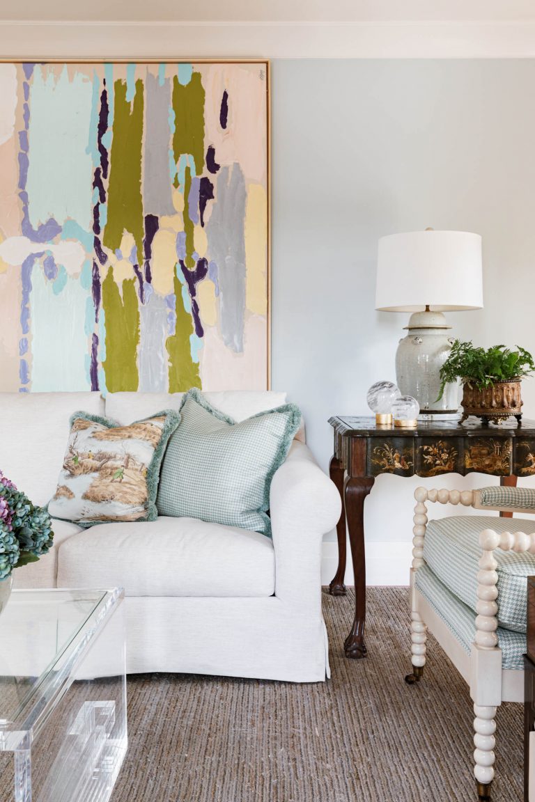 A living room features a white sofa with green and beige patterned cushions. A colorful abstract painting hangs above. Theres a glass table, a vintage wooden side table with a lamp and plant, and a striped upholstered chair. The carpet is woven in neutral tones.
