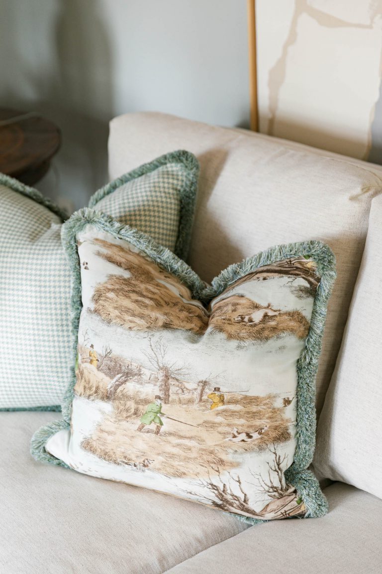 A beige couch features two decorative pillows. The front pillow has a pastoral scene with grass-green fringe, depicting people in a rustic landscape. The back pillow shows a light green and white checkered pattern. A wooden surface and soft wall colors complete the cozy setting.