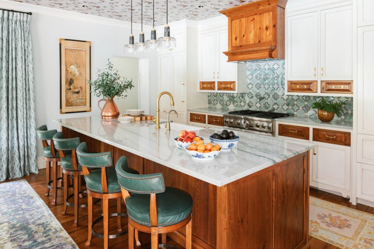 A spacious kitchen features a large wooden island with a marble countertop and green upholstered stools. A bowl of fruit is on the counter. White cabinets have wooden accents. The backsplash is blue and white tiles. A copper range hood and pendant lights add warmth. Curtains and artwork adorn the walls.