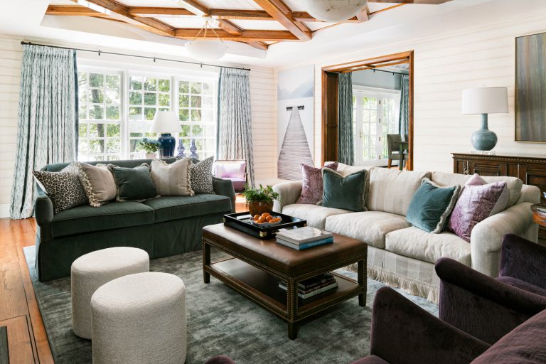 A cozy living room with plush sofas adorned with various pillows, a wooden coffee table holding books and a bowl of oranges, and two round white poufs. Large windows with patterned curtains let in natural light. The room features wooden accents and a carpet on the hardwood floor.