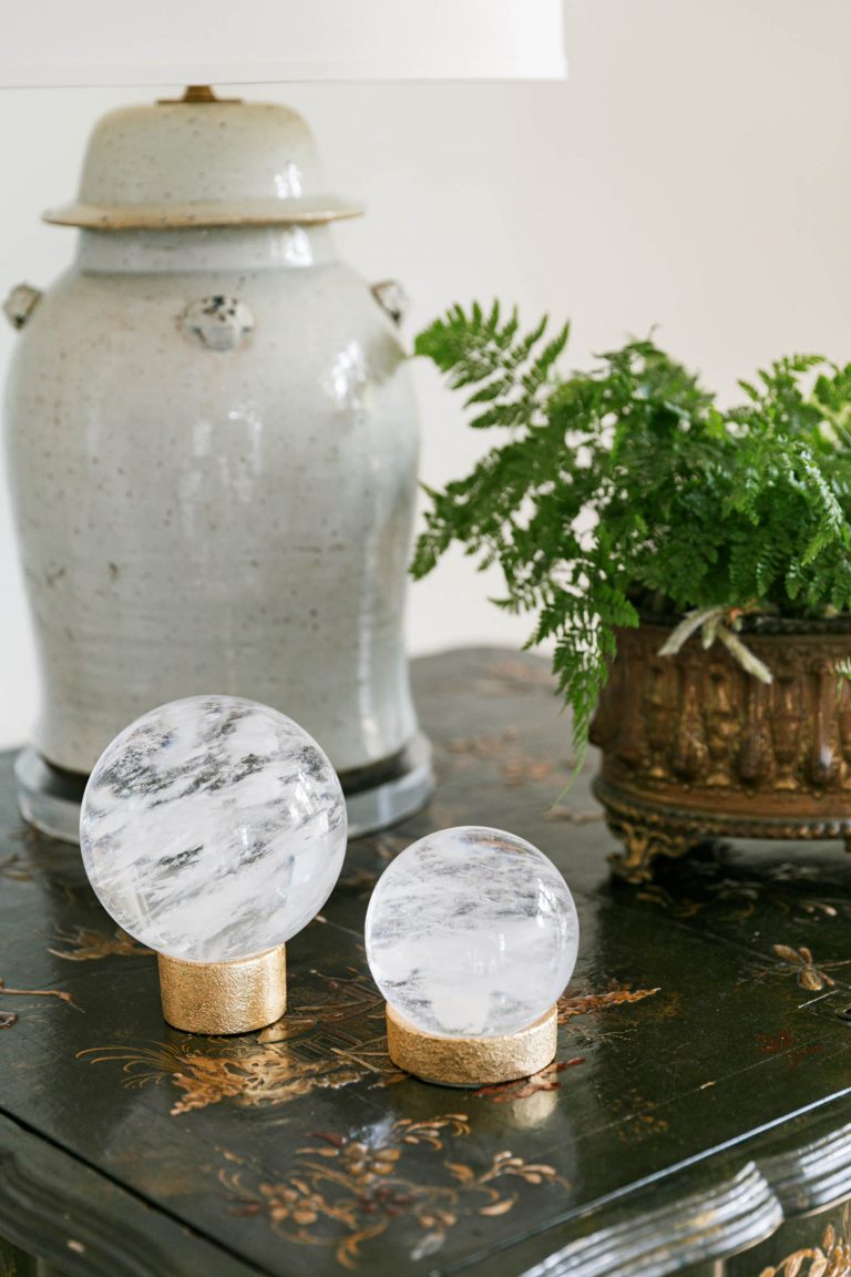 A decorative table displays two crystal spheres on gold bases. Behind them, a white ceramic lamp is positioned to the left, and a lush green fern sits in an ornate pot to the right. The table has a dark, glossy finish with intricate floral designs.