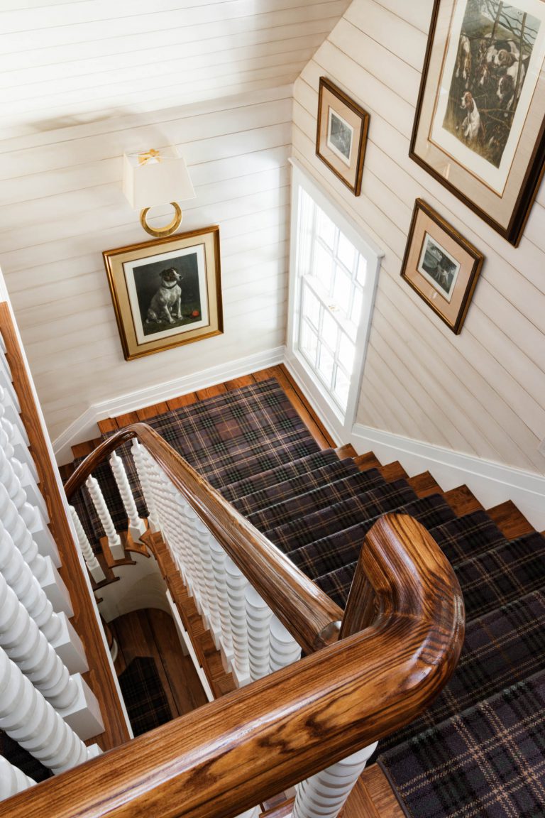 A wooden staircase with plaid carpet leads downward. The walls are lined with framed artwork, including a portrait and animal-themed pieces. A window on the right allows natural light in. The handrail is polished wood, and the wall has horizontal white paneling.