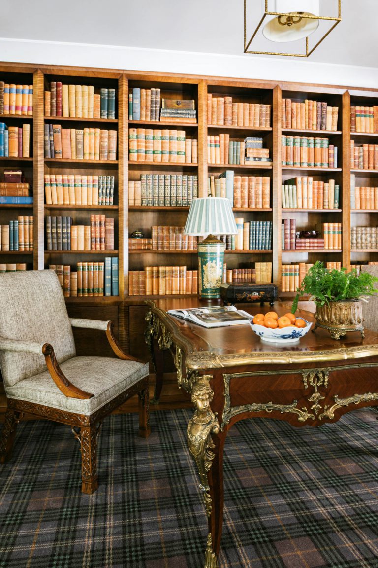 A classic library room with a bookshelf filled with leather-bound books. A wooden desk with ornate gold detailing holds a lamp, a bowl of oranges, and a potted plant. An upholstered armchair with wooden arms is nearby. The carpet has a plaid pattern in muted tones.