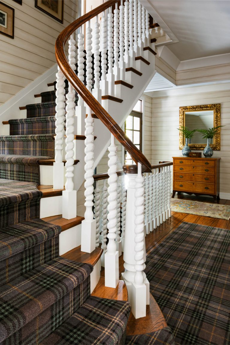 A staircase with dark plaid carpet and white spindle railings curves upward in a well-lit hallway. The walls are white with horizontal wood panels. At the top, a window allows natural light. A wooden dresser with a mirror and decorative items stands against the wall.