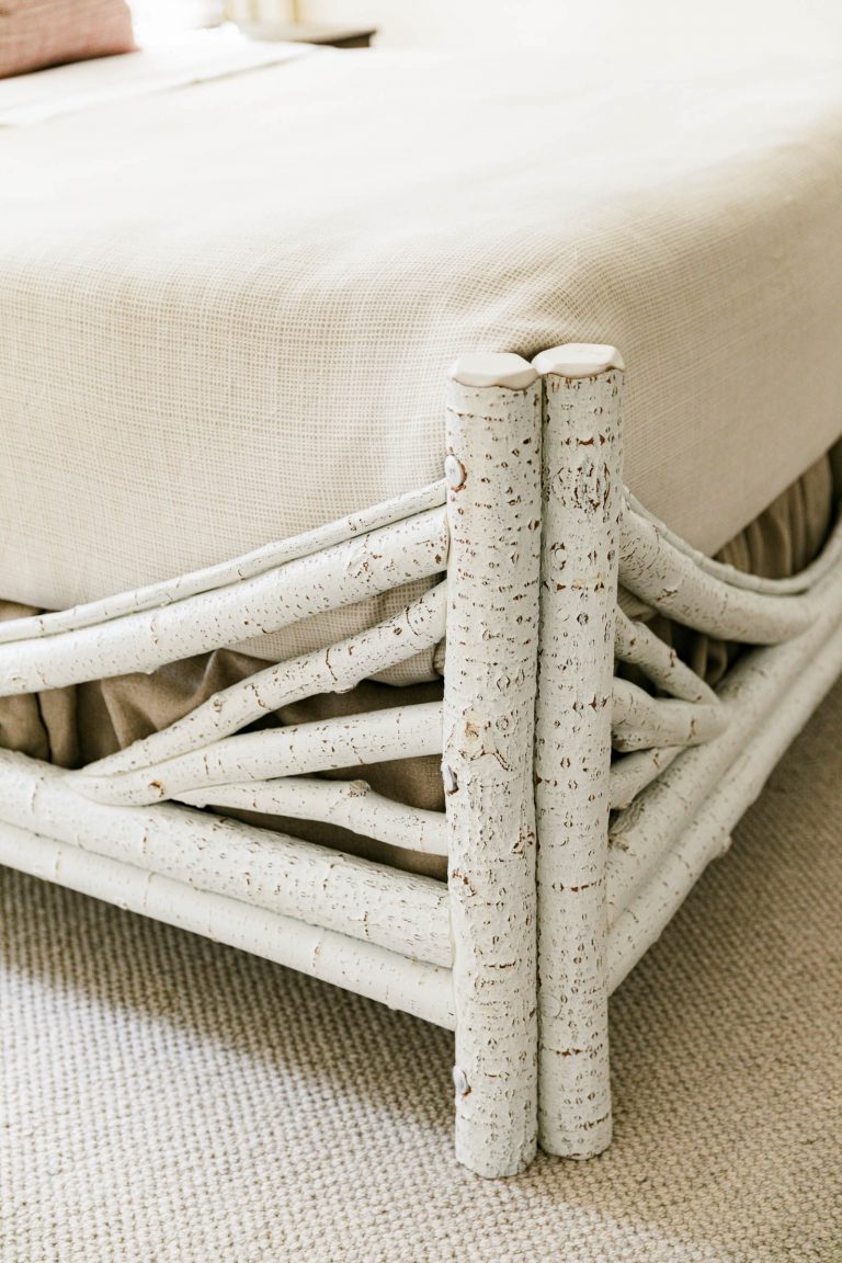 Close-up of a bed corner with a rustic wooden frame made of birch logs, displaying a natural, textured appearance. The bed is covered with a light beige, woven blanket, and rests on a neutral-colored, carpeted floor, creating a cozy and natural aesthetic.