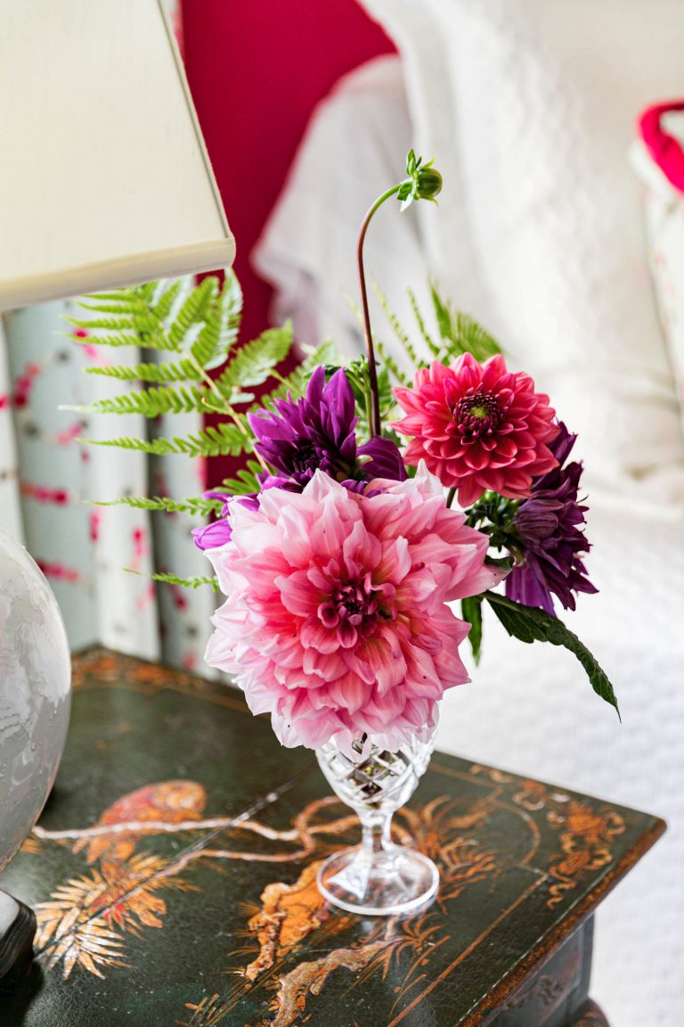 A crystal vase on a decorative table holds a floral arrangement with vibrant pink and purple dahlias, accented by green ferns. The background features a white bed and colorful curtains, creating a cozy and elegant bedroom setting.