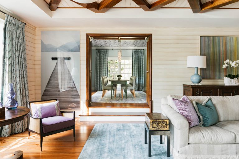 A cozy living room with wooden floors, featuring a cream sectional sofa, purple and blue pillows, a light blue rug, and a decorative side table. A large wall mirror reflects a dining area. Curtains with a subtle pattern frame a door, and a large print of a dock on a lake adorns the wall.
