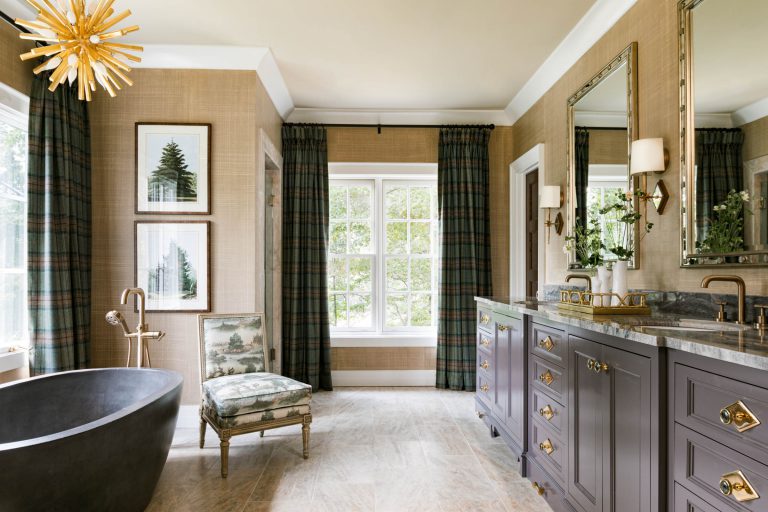 A luxurious bathroom features a black oval bathtub with a golden faucet, a cushioned chair with a green pattern, and double gray vanities with ornate mirrors and lamps. The room has large windows with plaid curtains, beige walls, and framed nature art. A starburst chandelier lights the space.