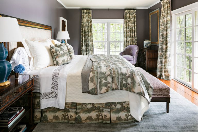A bedroom featuring a large bed with white and floral-patterned bedding, flanked by wooden nightstands with blue lamps. A purple armchair sits by the window with floral curtains. A bench is at the foot of the bed, and hardwood floors partially covered by a gray rug.