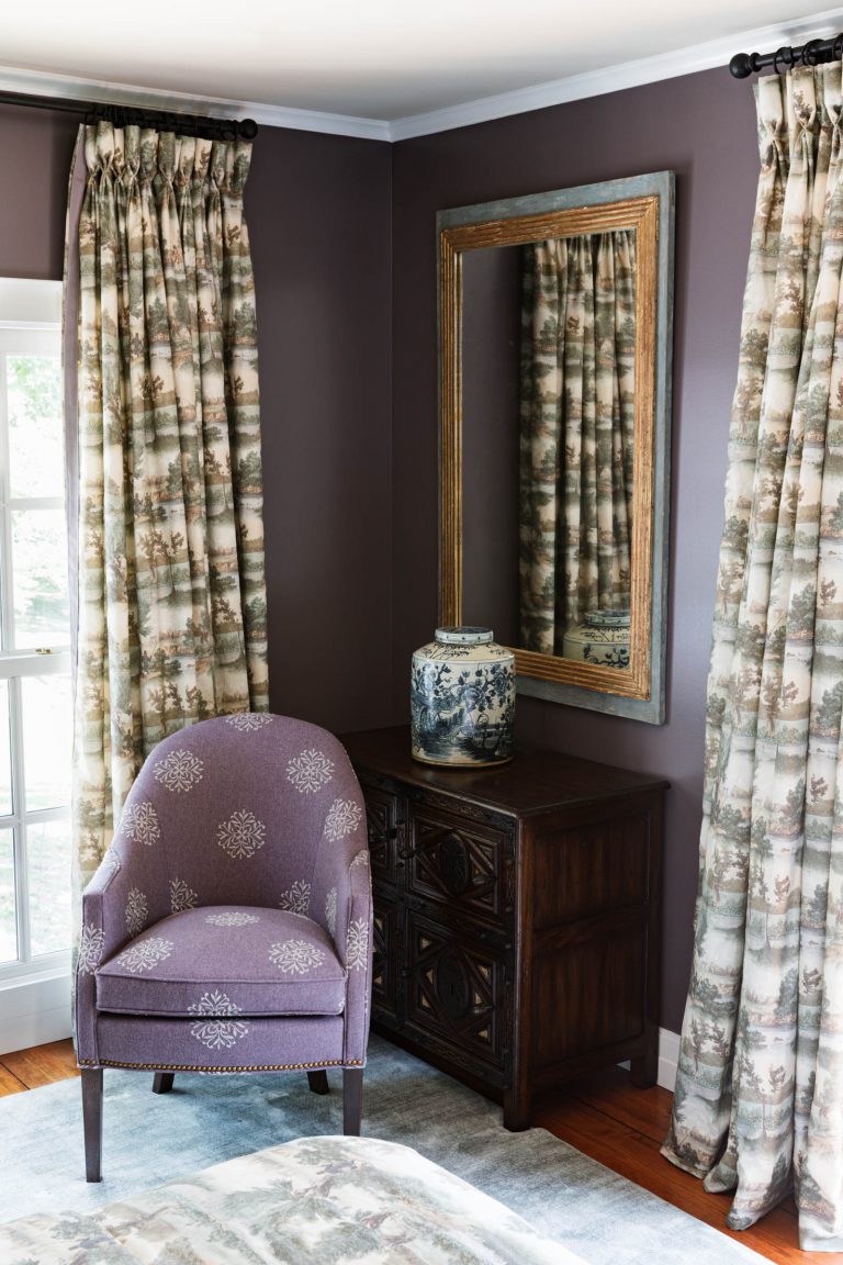 A cozy room corner with deep purple walls features a purple upholstered armchair with floral patterns. A dark wooden dresser holds a blue and white ceramic vase. A large mirror in a wooden frame reflects the room, while patterned curtains hang beside a window. A patterned rug is on the floor.