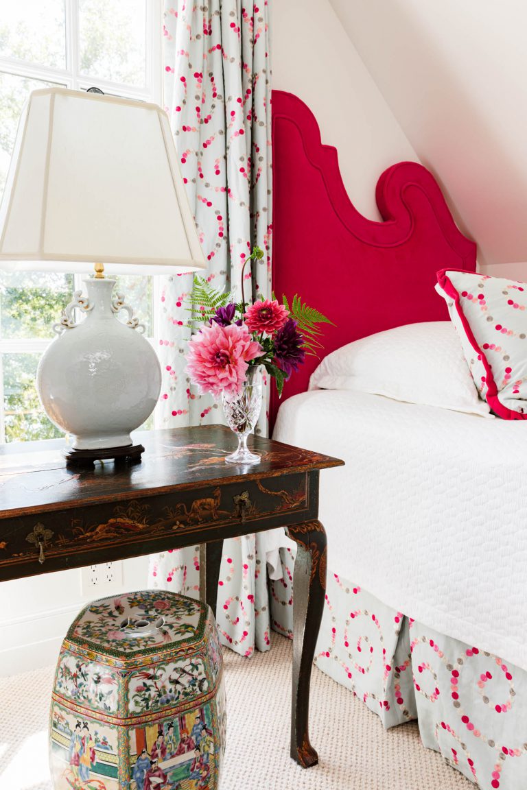 A bright bedroom featuring a bed with a vivid pink, scalloped headboard and white bedding. A dark wooden side table with decorative details holds a ceramic lamp and a vase of vibrant flowers. A colorful floral stool is underneath. The curtains are white with pink patterns, matching the bed skirt.