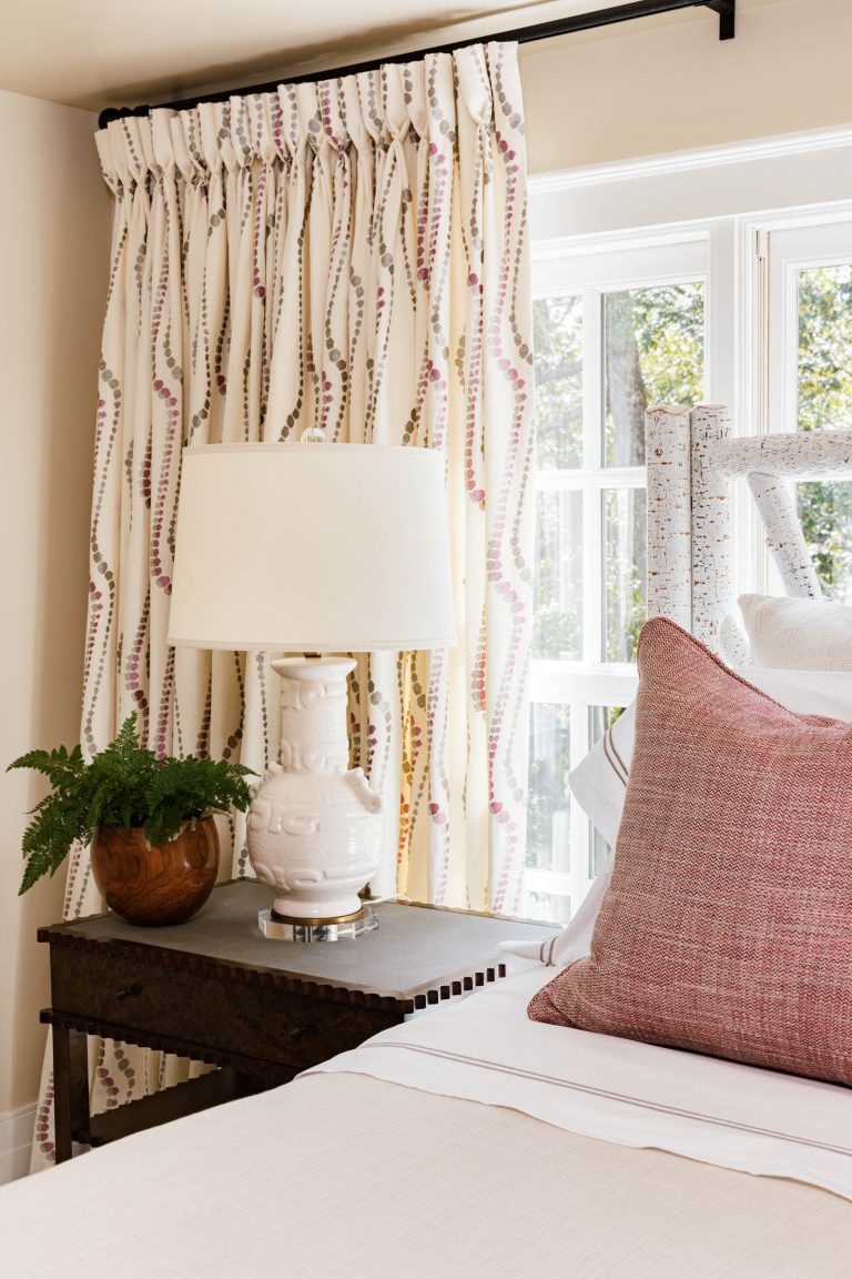 A cozy bedroom corner featuring a white bed with a textured pink pillow. A dark wooden side table holds a white ceramic lamp and a brown pot with a fern. Curtains with pink and beige patterns hang beside a window, letting in natural light. The space exudes warmth and style.