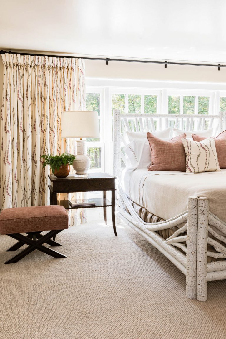 A cozy bedroom with a large white wooden bed, adorned with cream and blush pillows. A wooden side table holds a lamp and a potted plant. Light beige curtains with a subtle pattern cover a sunlit window. A brown upholstered stool sits nearby on a beige carpet.