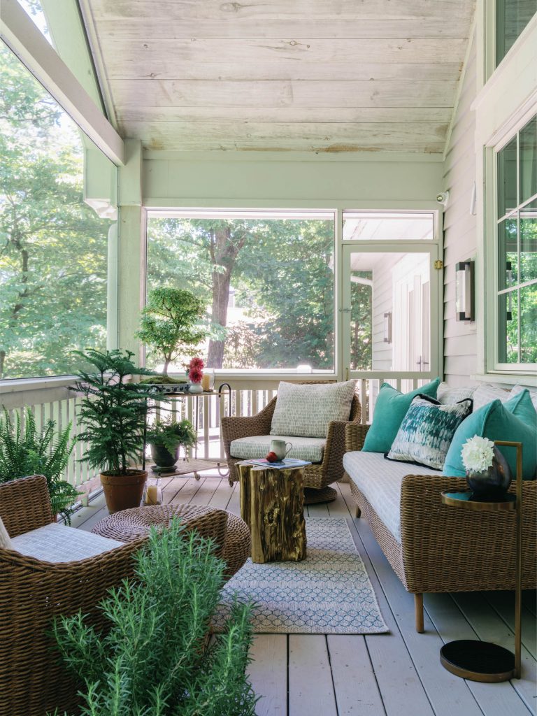 A cozy patio with wicker furniture, including a sofa and two chairs, adorned with teal and white cushions. A wooden stump serves as a coffee table. Potted plants surround the area, while large windows and a glass door offer a view of lush greenery outside. Light wood flooring enhances the space.
