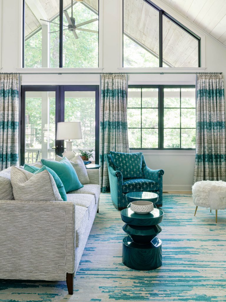 A modern living room with tall windows and blue-green decor. It features a grey sofa with teal cushions, a teal patterned armchair, and a fluffy white stool. A striped blue and white rug covers the floor, and a small round teal side table is topped with decorative bowls.