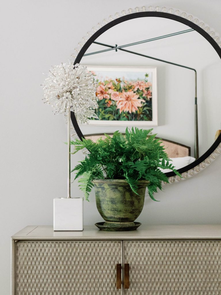 A stylish room features a circular mirror with a beaded edge above a textured cabinet. On the cabinet, theres a tall, decorative white sculpture and a green fern in a ceramic pot. A floral painting with pink flowers is reflected in the mirror, adding color to the neutral space.