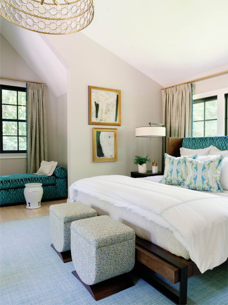 A cozy bedroom features a large bed with white and blue bedding, two patterned ottomans at its foot, and a textured blue headboard. The walls display abstract art. A window with patterned curtains illuminates the room, which includes a blue chaise lounge and a floor lamp. A modern light fixture hangs from the ceiling.
