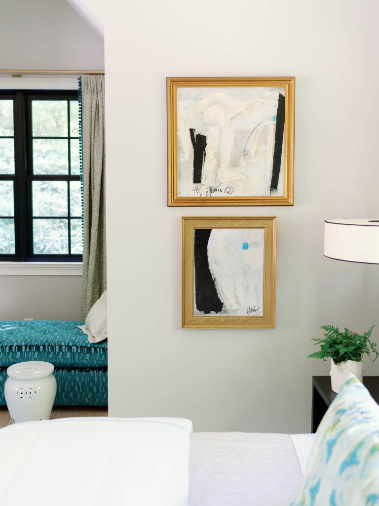 Interior scene with two abstract paintings in gold frames on a light wall. Below is a potted plant on a white surface. A window with black frames and blue curtains is visible, alongside a turquoise upholstered bench and a small white ceramic stool. A portion of a white lampshade is seen.