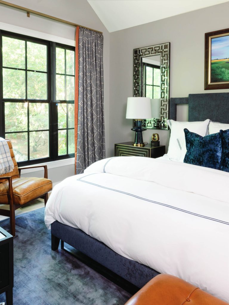 Bedroom with a large bed featuring white linens and dark blue pillows. A window with patterned gray curtains allows natural light in. Beside the bed is a nightstand with a table lamp and framed mirror above. A leather chair sits near the window. A framed landscape painting hangs on the wall.