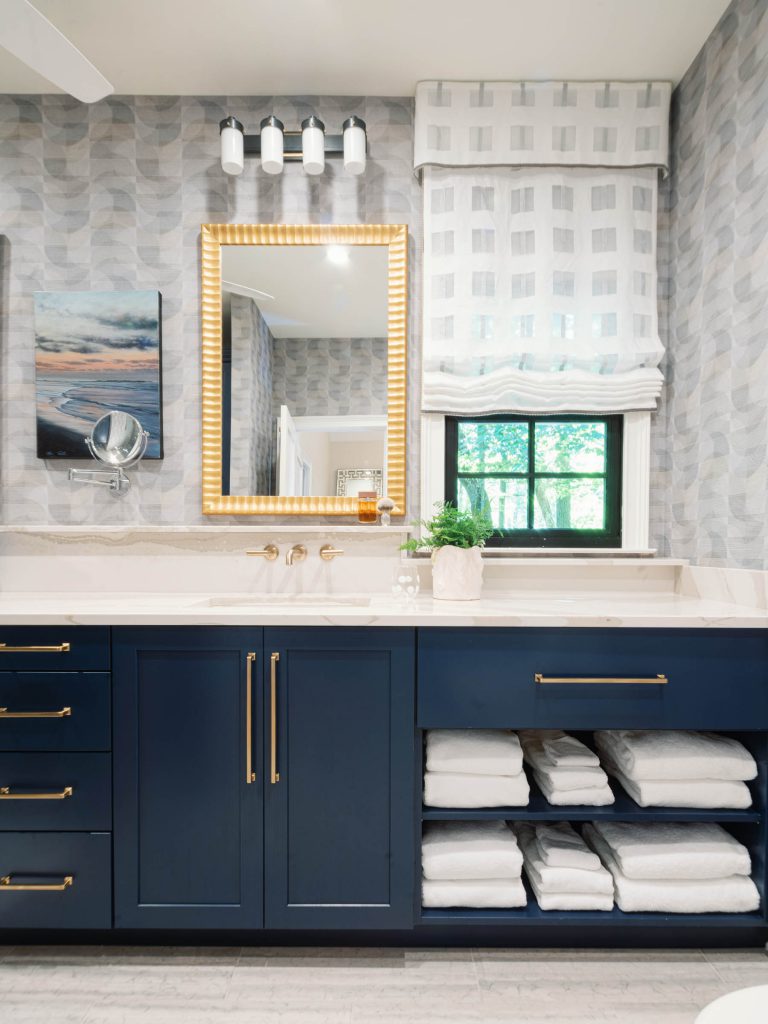 A modern bathroom features a navy blue vanity with gold hardware, a lighted mirror, and a white countertop. The wall has patterned wallpaper, and a window with a white roman shade lets in natural light. Towels are neatly stacked on open shelves below the counter.