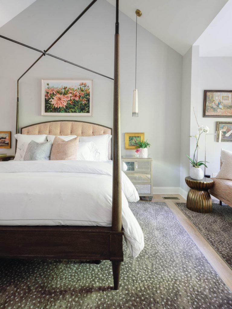 A cozy bedroom featuring a canopy bed with white bedding and pastel pillows. A nightstand with a lamp and framed art is beside the bed. There is a large floral painting above the headboard. A textured rug covers the floor, and an orchid plant is placed on a round side table.