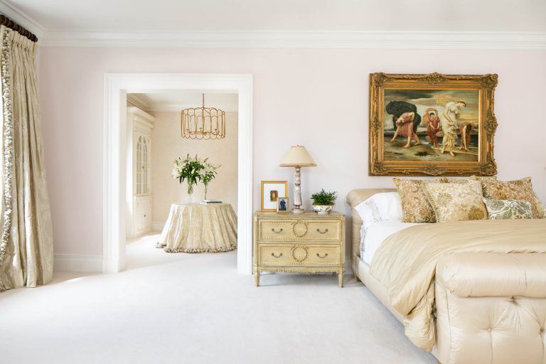 Elegant bedroom with light beige walls and bedding. A large framed painting above the bed features classical figures. A vintage nightstand holds a lamp and greenery. Through the open doorway, a small round table with a floral arrangement and a decorative chandelier are visible.