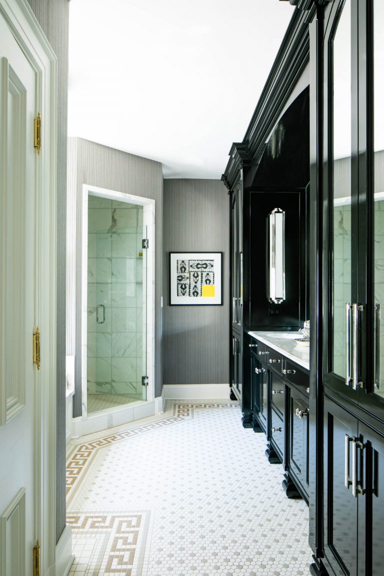 Elegant bathroom with black cabinetry and a white marble countertop. A glass-enclosed shower with marble tiles is on the left. The floor features mosaic tiles with a geometric border. A framed artwork with a splash of yellow hangs on the light gray wall.