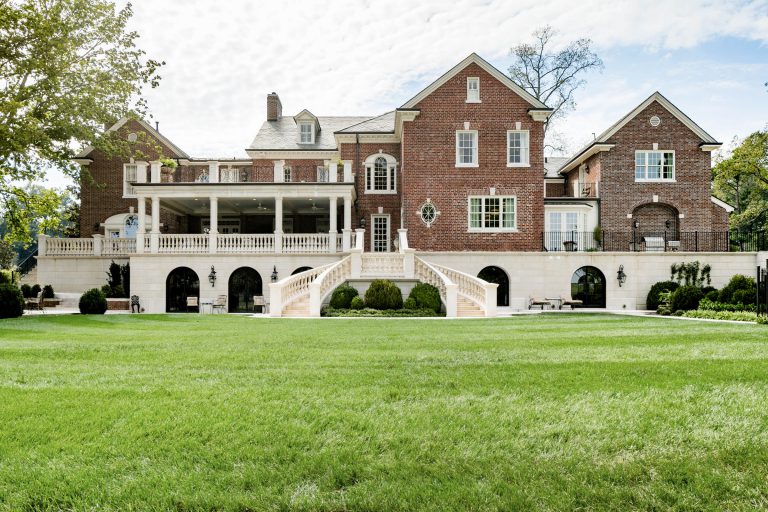 A large, elegant brick mansion with multiple stories, featuring white accents, arched windows, and a grand staircase leading to a landscaped lawn. The property is surrounded by greenery and a partially cloudy sky in the background.