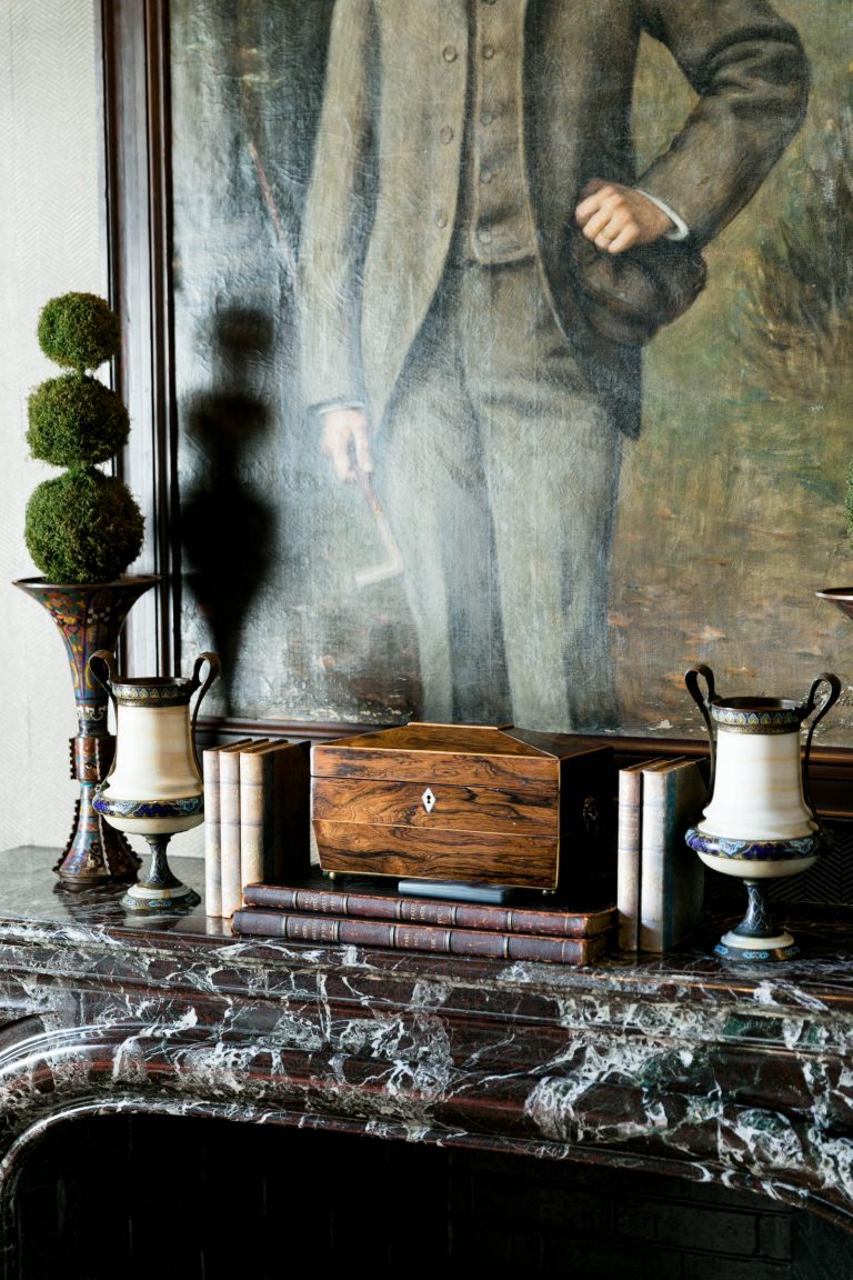 A luxurious interior with a marble fireplace mantel adorned with two urn-shaped vases and stacked books. A wooden box rests centrally. Above, a large painting depicts a person in a suit with a hand on their hip. Two topiary spheres add greenery to the elegant setting.