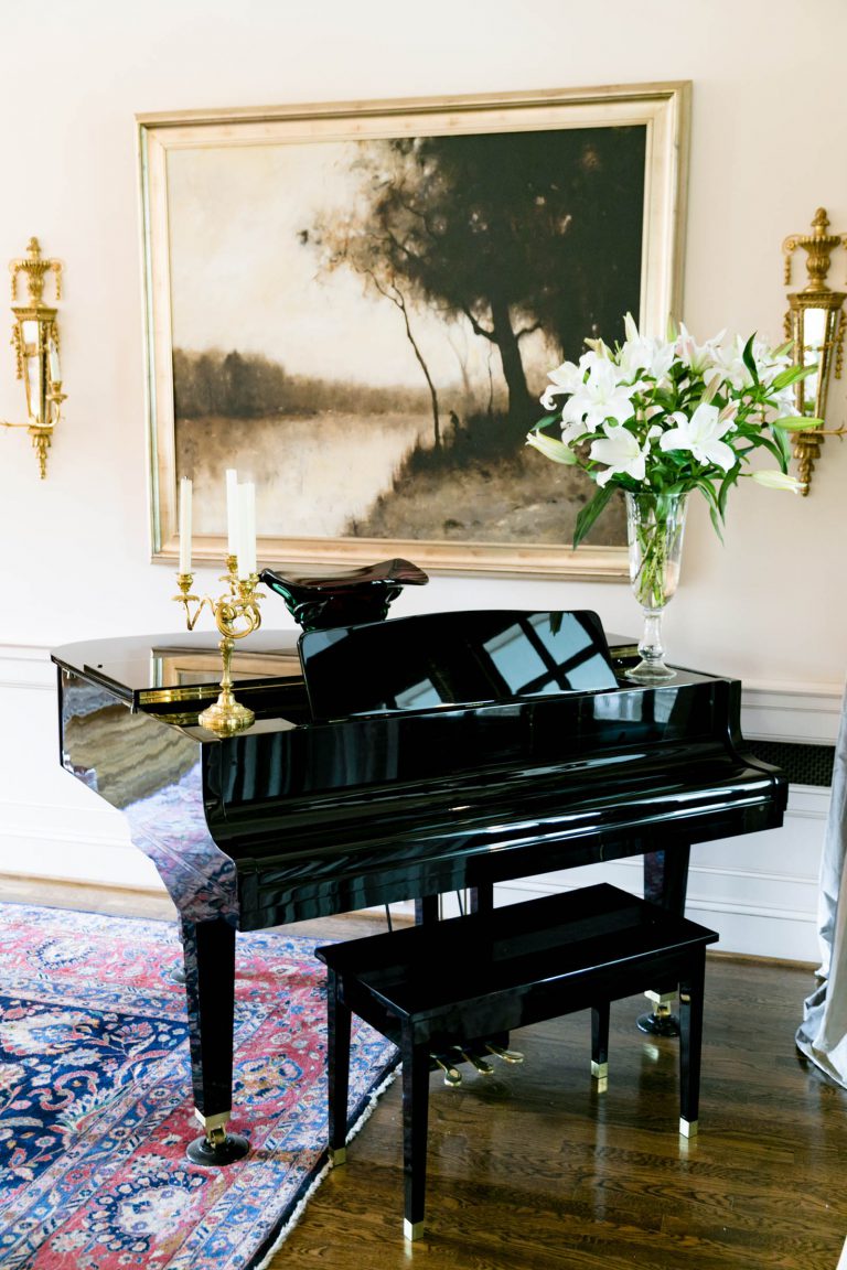 A polished black grand piano with a matching bench is set on a wooden floor adorned with a colorful patterned rug. It features a brass candle holder and a vase of white lilies. A landscape painting and ornate wall sconces decorate the background wall, enhancing the elegant setting.