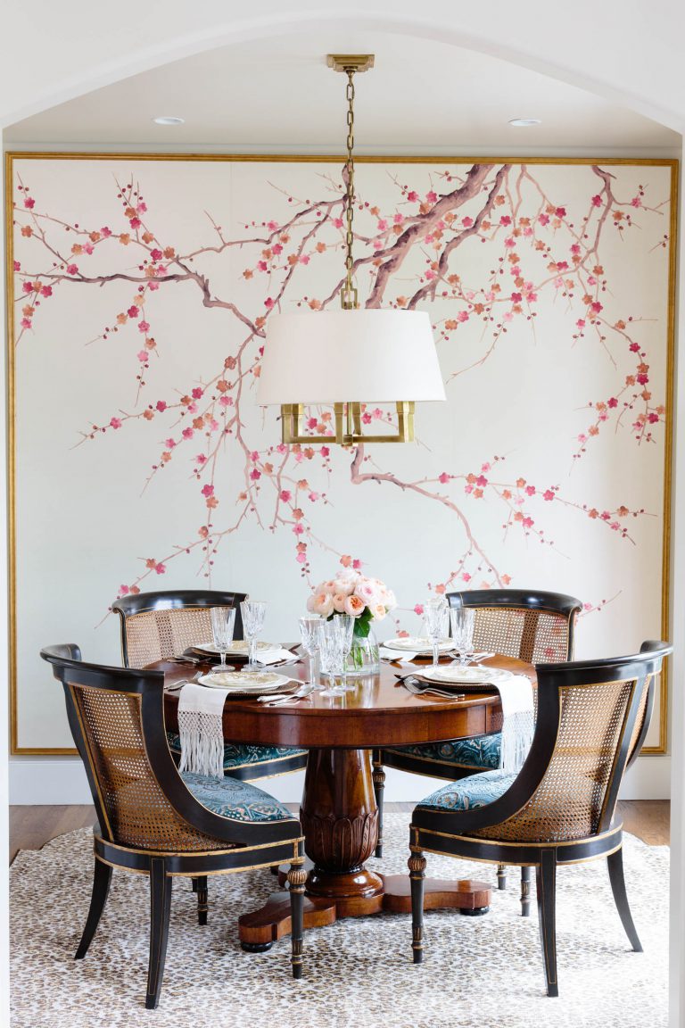 A dining room featuring a round wooden table set for four with glassware and a floral centerpiece. Four cane-back chairs surround it. The backdrop wall has a mural of pink cherry blossoms. A large, elegant pendant light hangs above, and a neutral rug lies on the floor.