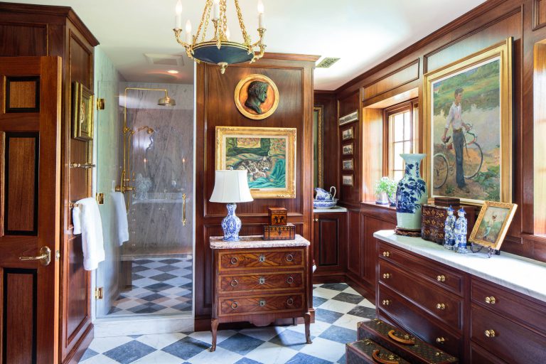 Elegant bathroom with rich wooden paneling and a black-and-white checkered floor. It features ornate chandeliers, a freestanding wooden chest with a lamp, and framed artwork. A glass-enclosed shower is visible on the left, and large windows provide natural light. Blue vases decorate surfaces.