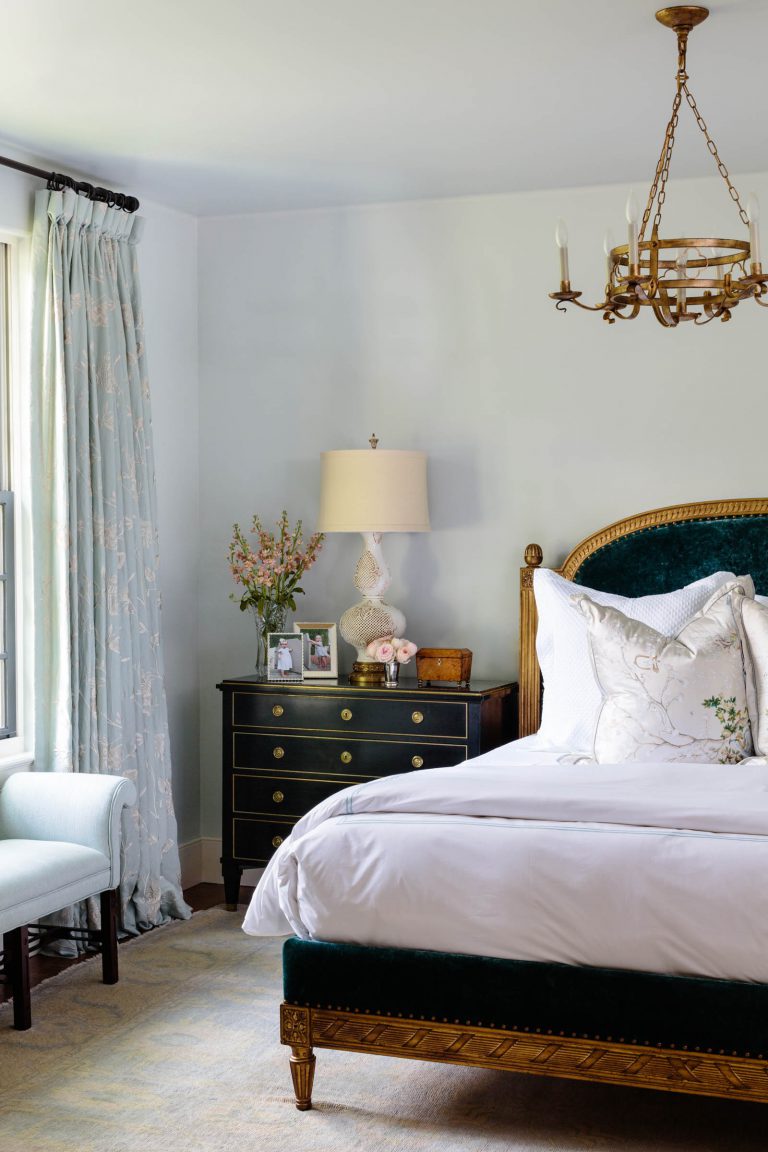 A cozy bedroom featuring a plush bed with white linens and a green upholstered headboard. Beside the bed is a dark wooden nightstand with a lamp, flowers, and framed photos. A window with light blue curtains and a light blue upholstered bench are to the left. A chandelier hangs from the ceiling.