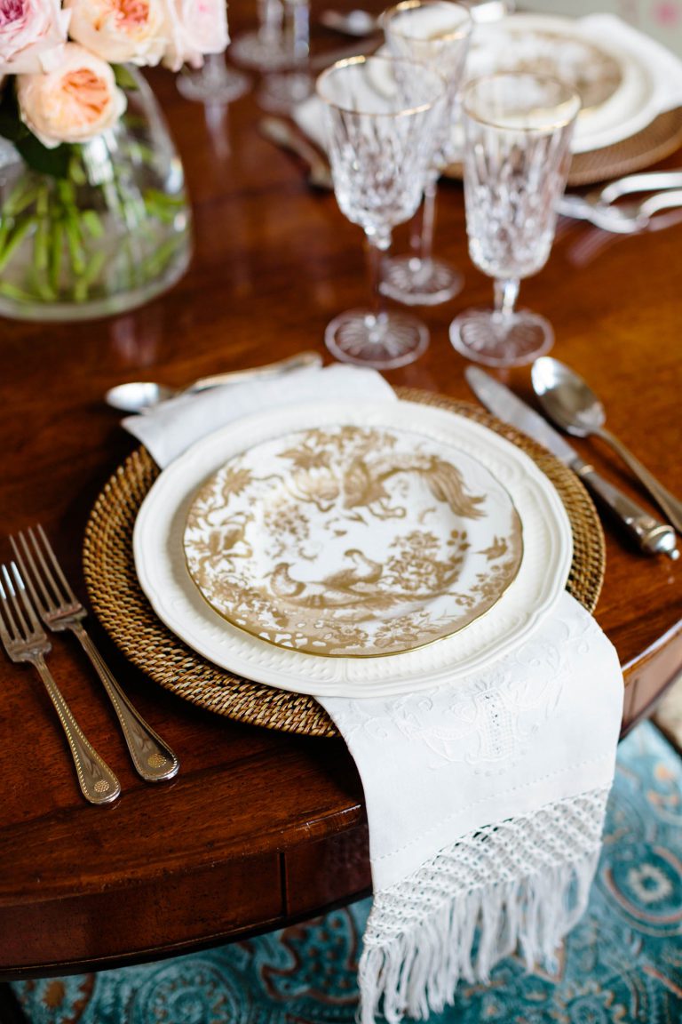 A close-up of an elegant table setting on a wooden table. A decorative gold and white plate rests on a woven charger, with a white fringed napkin beneath. Crystal glassware and silver cutlery are neatly arranged. A floral centerpiece with pink roses is partially visible in the background.
