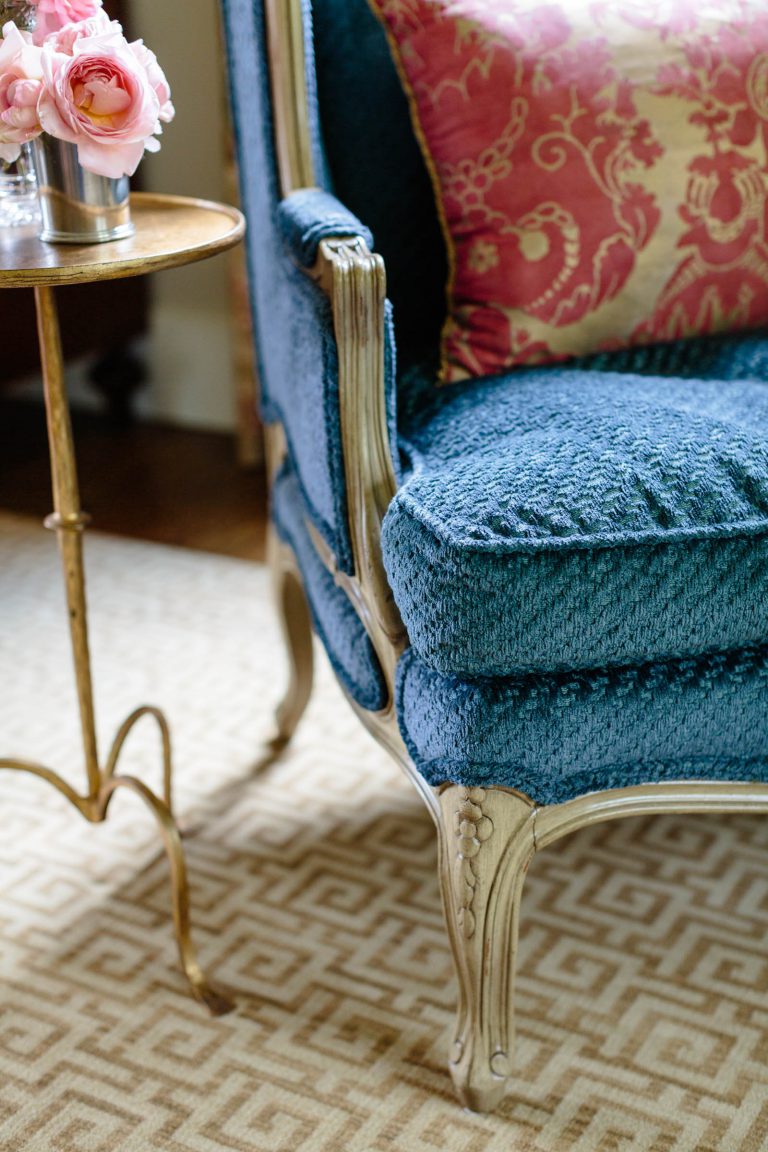 A luxurious blue upholstered armchair with ornate silver wooden legs and armrests is positioned next to a small round table. The table holds a vase of pink roses. In the background, a pink decorative pillow with a floral pattern complements the classic setting. A patterned rug is partially visible.