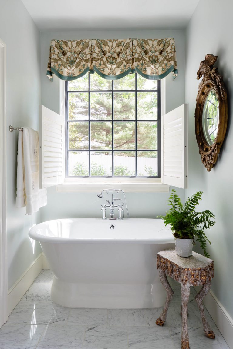 A bathroom featuring a white freestanding bathtub beneath a large window with shutters. The window has a floral valance and looks out onto trees. A rustic wooden side table holds a potted fern. A circular ornate mirror hangs on the wall, and a towel is on a rack nearby.