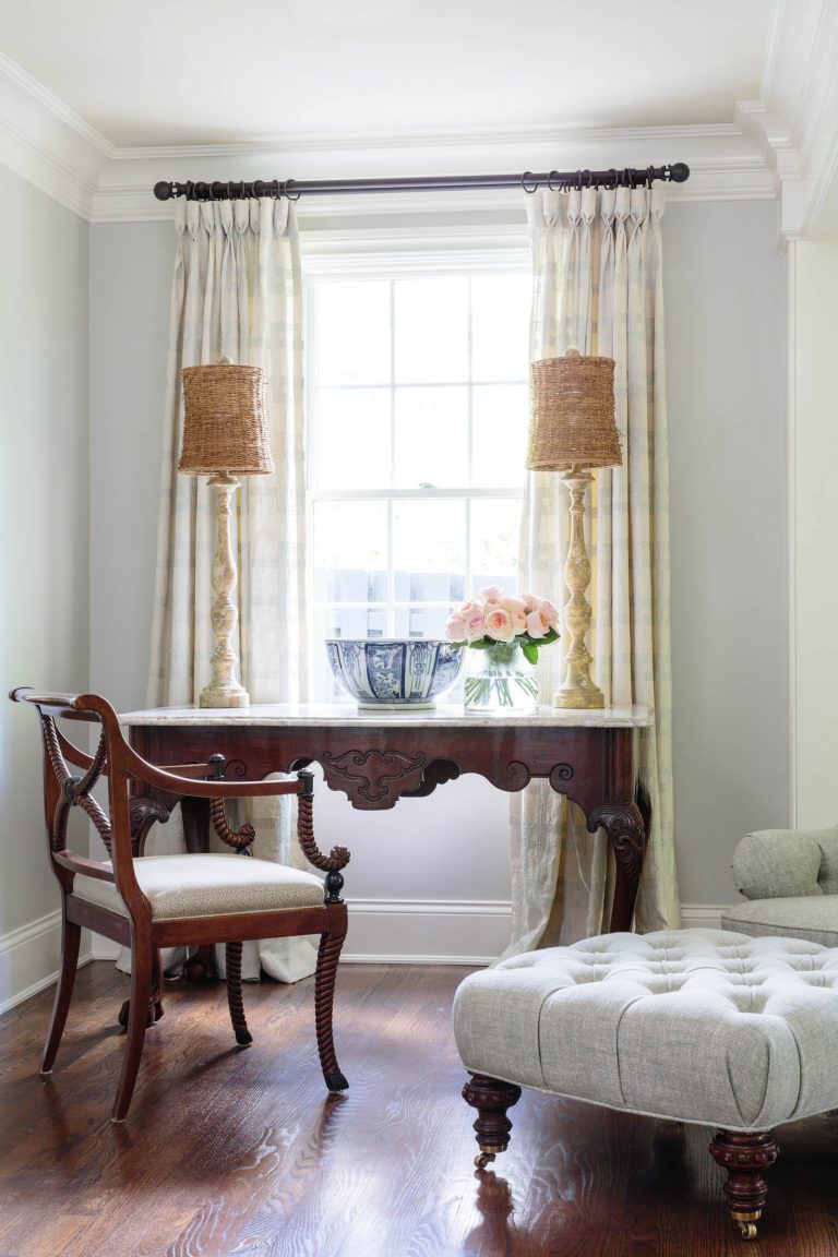 A classic room features a wooden writing desk with ornate carvings. On the desk are two tall wicker lamps and a blue vase holding pink roses. A wooden chair with a cushioned seat is nearby. A tufted footstool is on the dark hardwood floor. Light curtains frame the window behind.