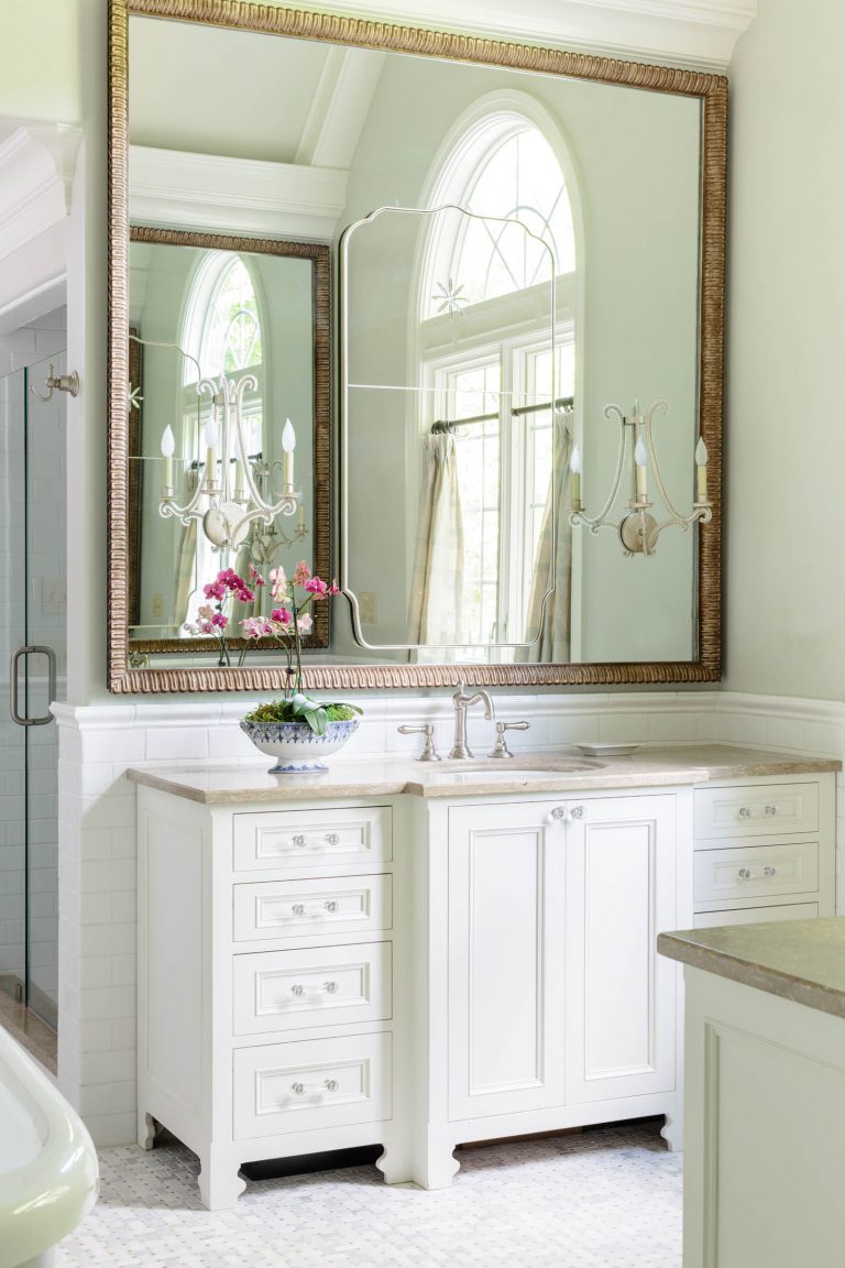 A traditional bathroom features a large ornate mirror above a white vanity with brass fixtures. The vanity has drawers and cabinets. A sconce with candles is reflected in the mirror alongside an arched window. A bowl with pink flowers adds a touch of color to the predominantly white decor.